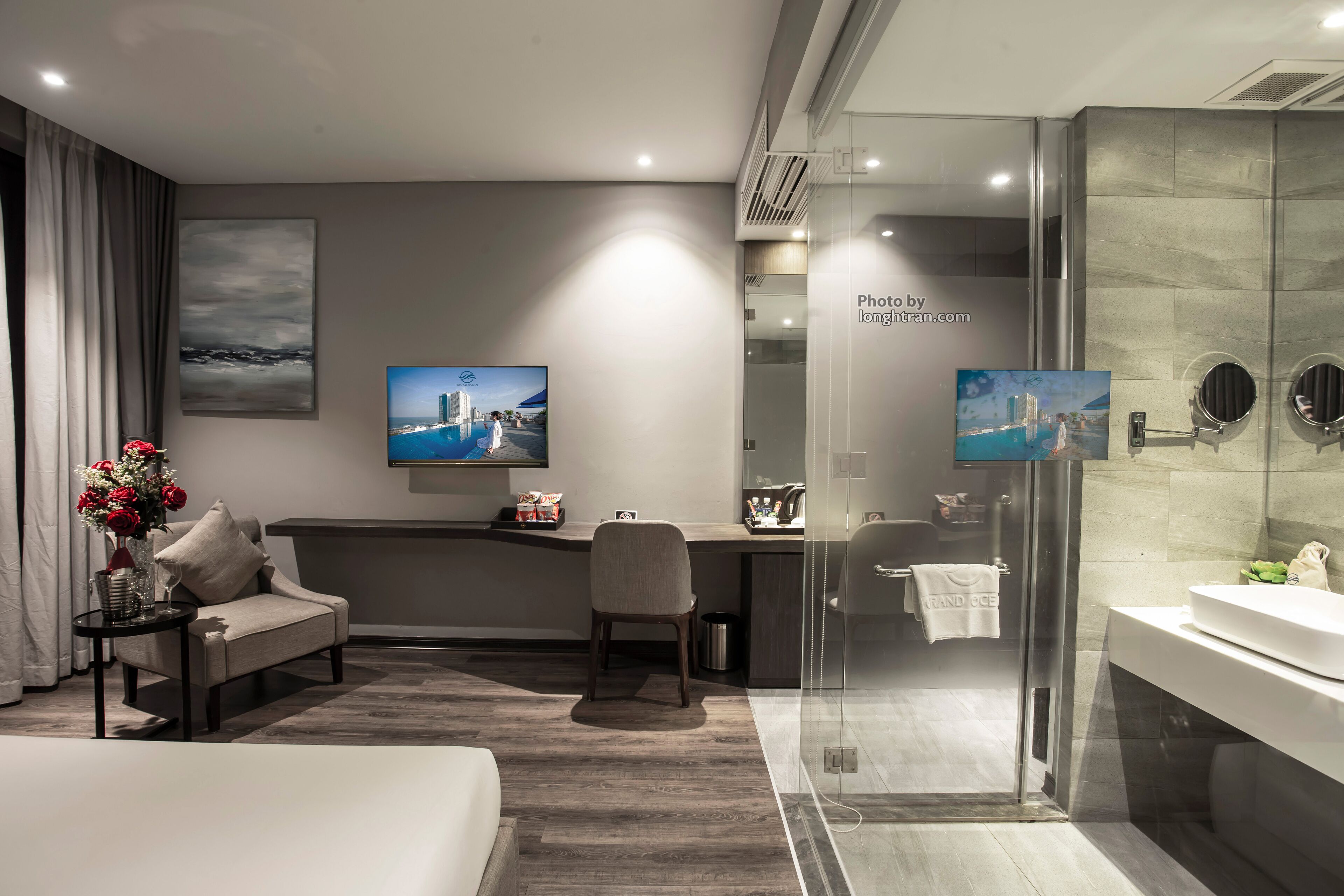 A modern hotel room with a bed, a desk, a chair, a TV, and a bathroom with a glass shower enclosure.