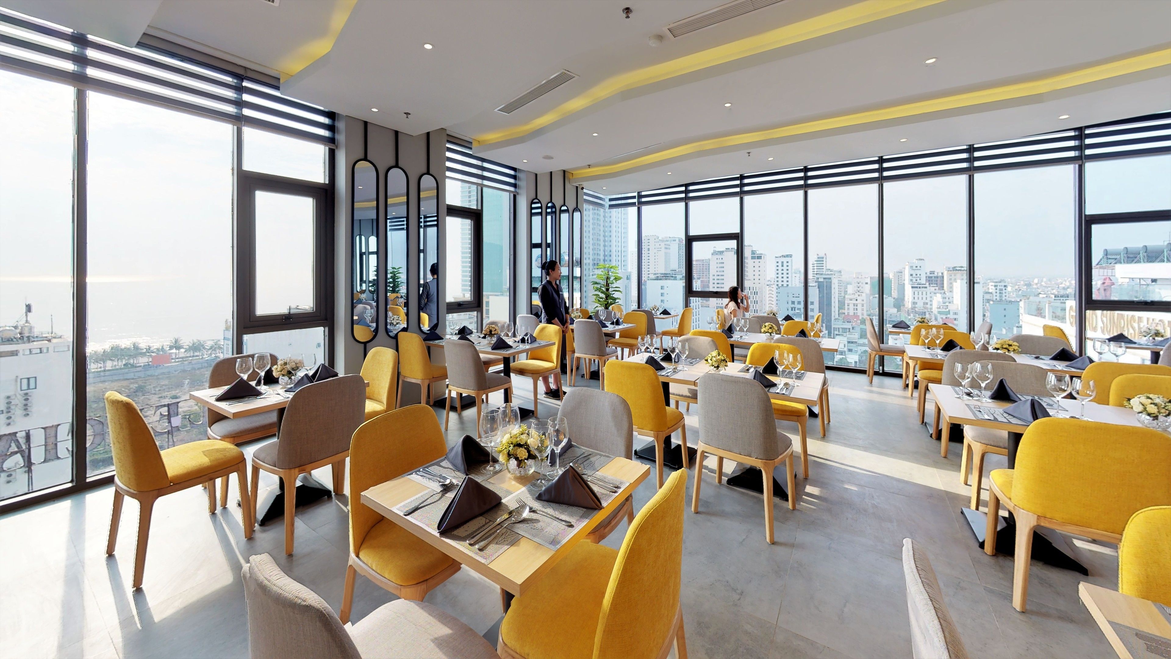 A modern dining area with yellow chairs and tables, large windows offering a city view, and a clean, contemporary design.