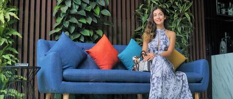 A woman sitting on a blue sofa with colorful cushions, in front of a wooden wall with vertical slats and green plants.