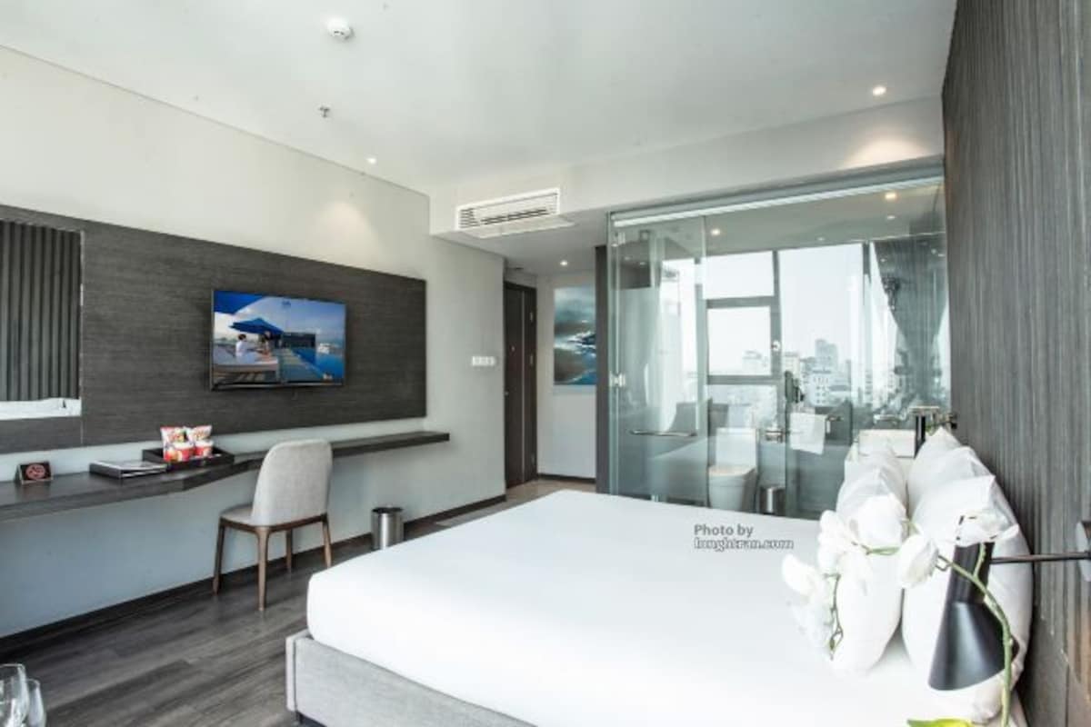 Una habitación de hotel moderna con una cama grande, un escritorio con una silla, un televisor y una puerta de vidrio que da a un balcón con vistas a la ciudad.