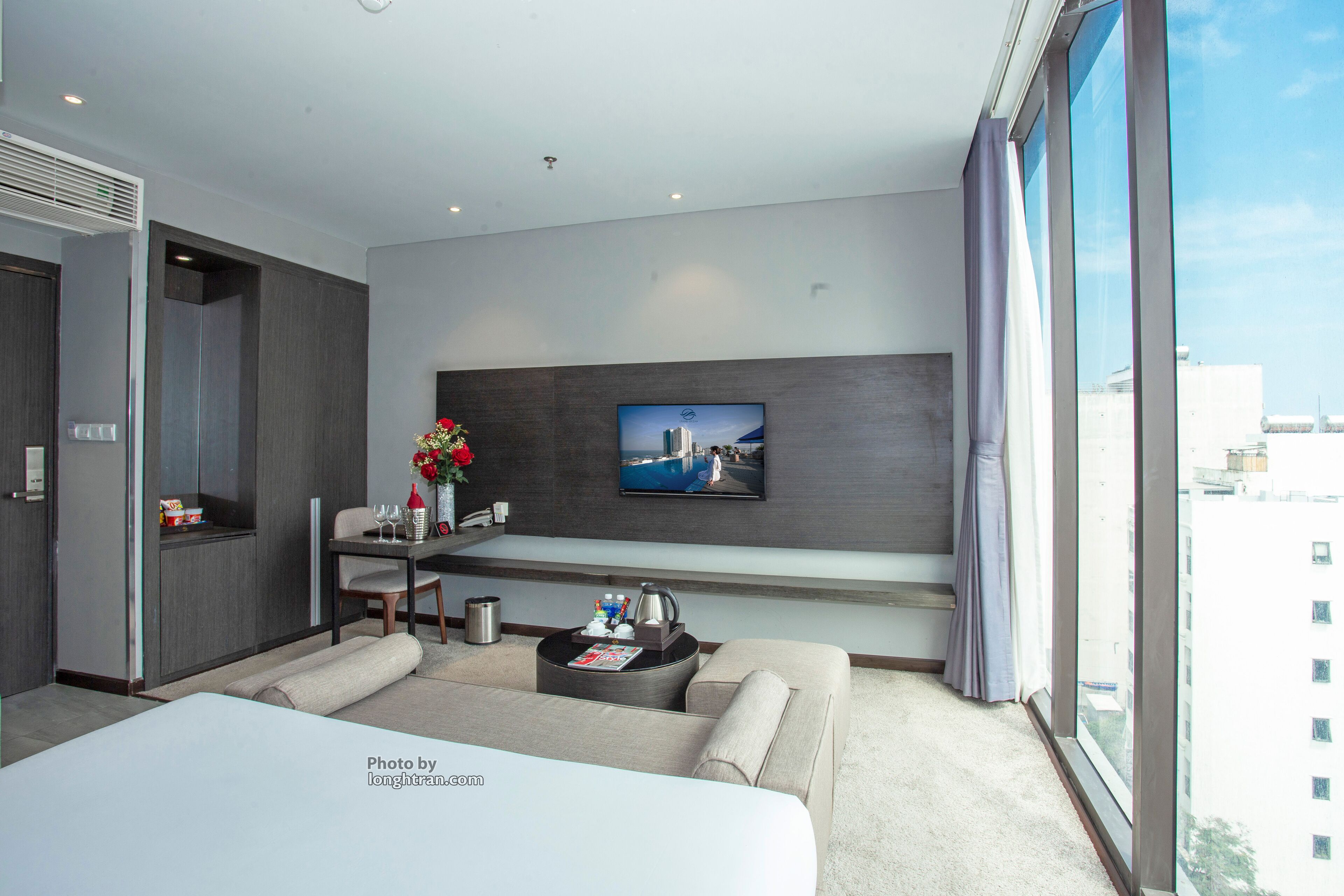 A modern hotel room with a large bed, a seating area with a coffee table, a TV mounted on the wall, and a window with a city view.