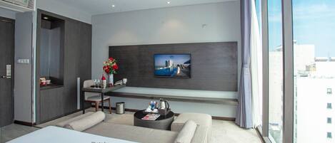 A modern hotel room with a large bed, a seating area with a coffee table, a TV mounted on the wall, and a window with a city view.