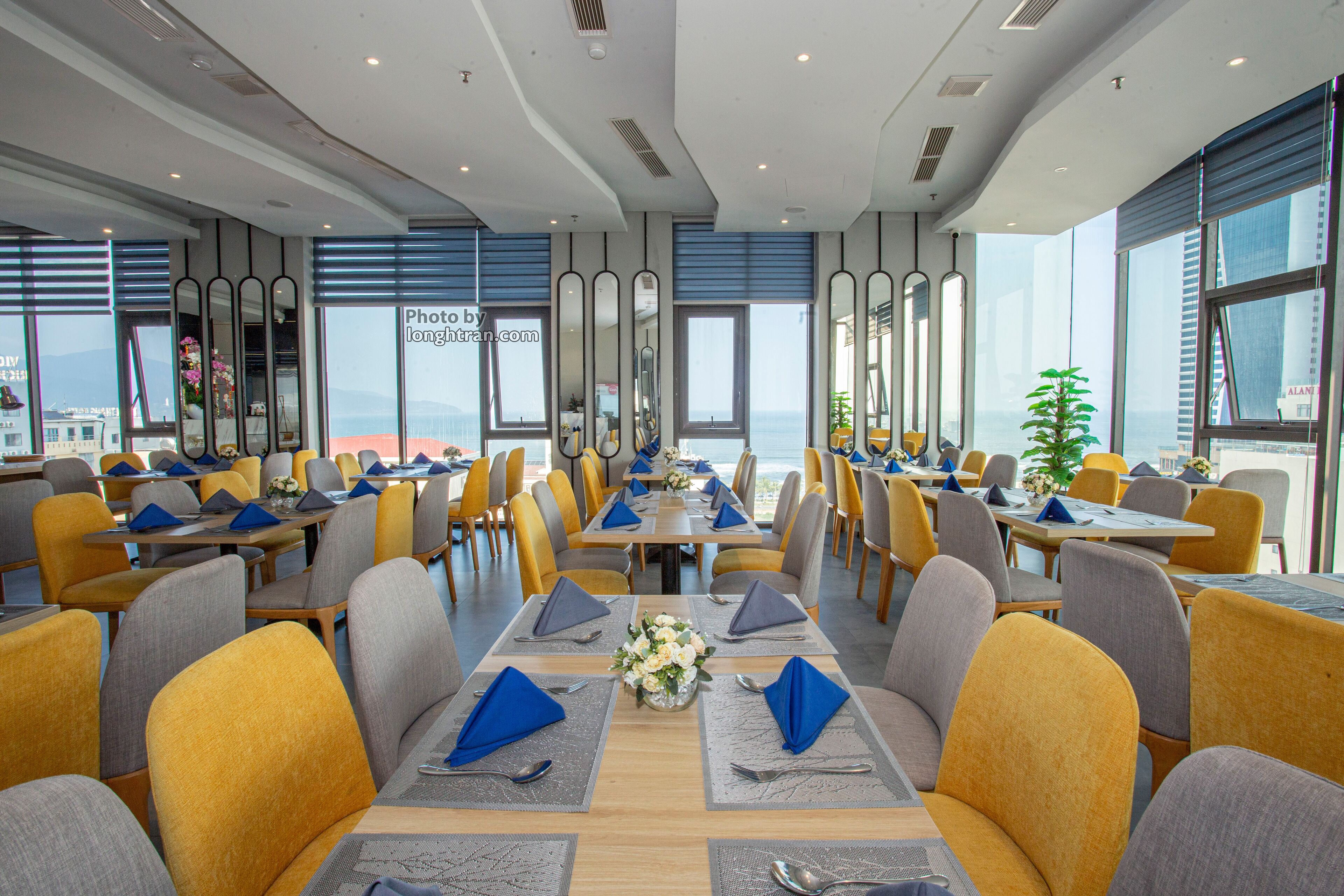 A modern dining area with tables set for four, yellow and gray chairs, and large windows offering a view of the sea.