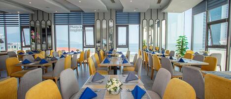 A modern dining area with tables set for four, yellow and gray chairs, and large windows offering a view of the sea.
