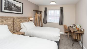 Habitación de hotel con dos camas, un escritorio con silla, una ventana con cortinas, un perchero y un cuadro en la pared.