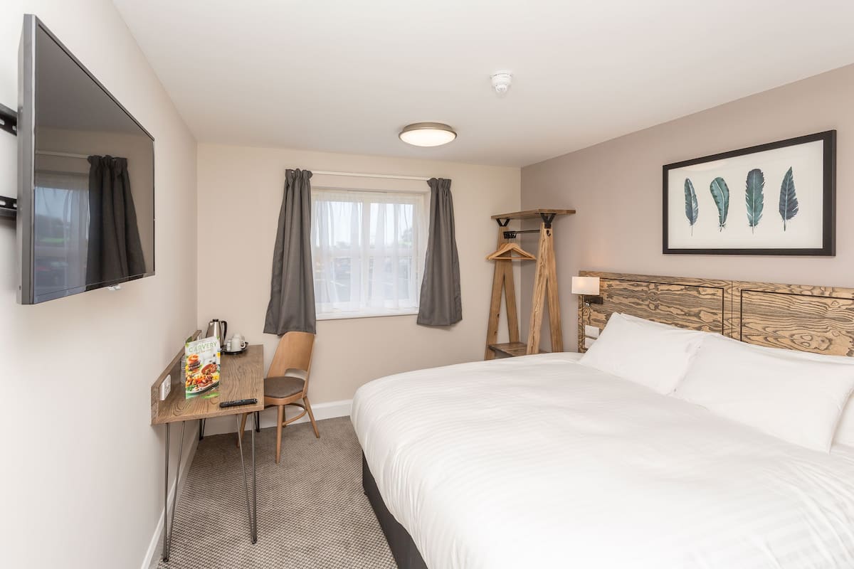 A hotel room with a bed, a desk with a chair, a television, a window with curtains, and a framed picture on the wall.