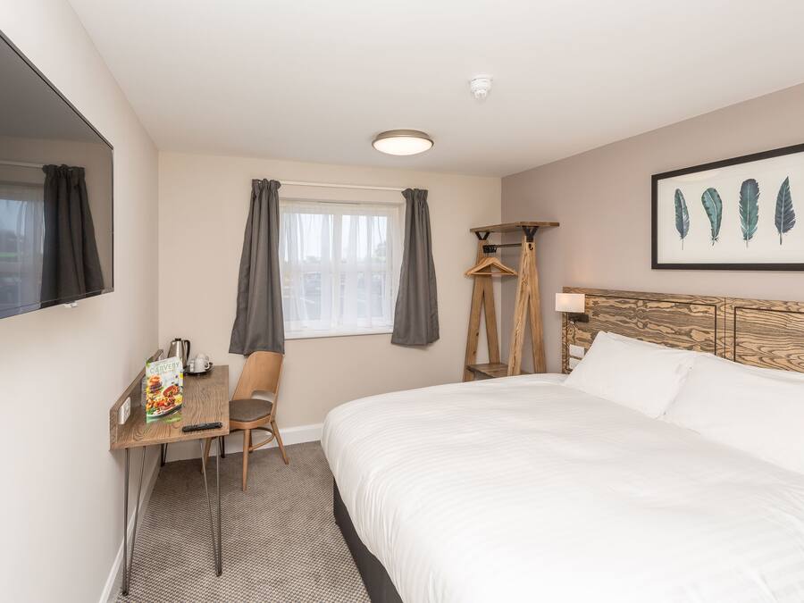 Habitación de hotel con cama, escritorio con silla, televisión, ventana con cortinas e imagen en un marco en la pared.