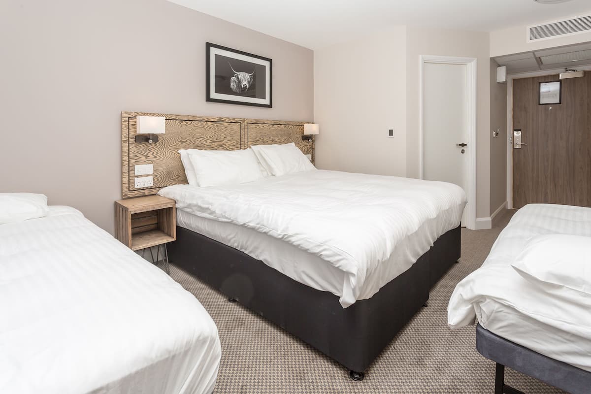 A hotel room with a large bed, two bedside tables, a headboard with a light fixture, a framed picture on the wall, and a door to the right.