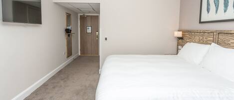 A hotel room with a large bed, a flat-screen TV mounted on the wall, a wooden headboard, and a framed artwork above the bed.