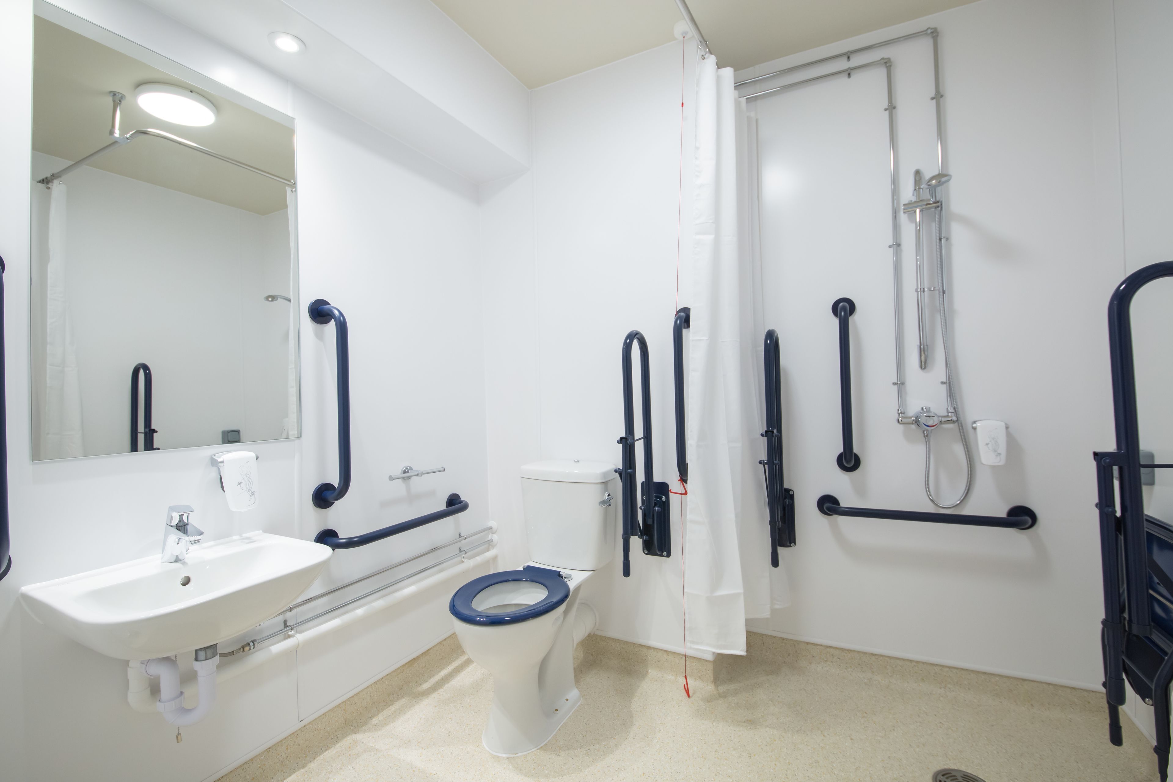 Un baño moderno con un lavamanos blanco, un inodoro con asiento azul, una ducha con cabezal de ducha manual y barras de apoyo.
