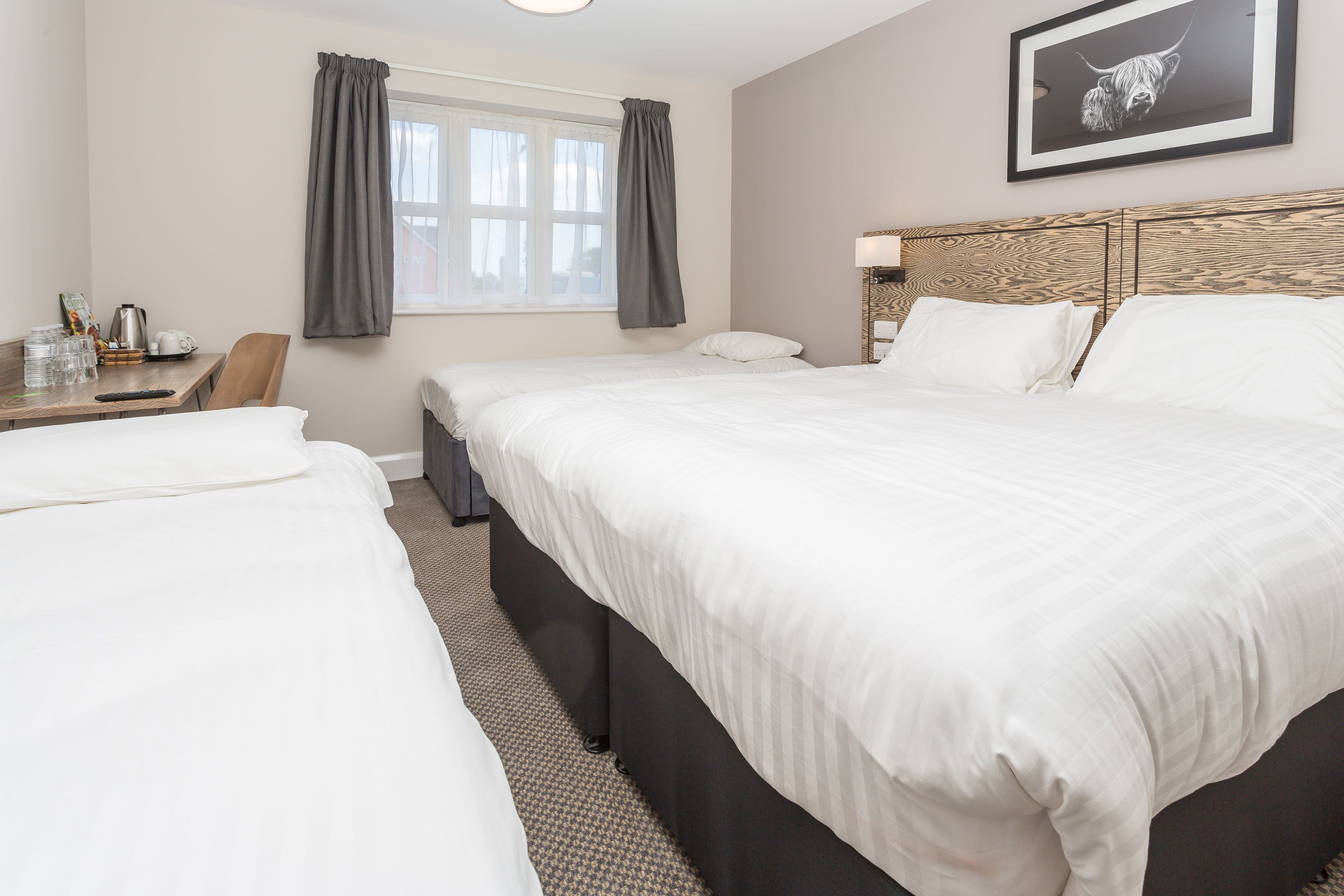 Une chambre d’hôtel avec deux lits simples, une tête de lit en bois, un bureau avec une chaise, une fenêtre avec des rideaux et un tableau encadré au mur.