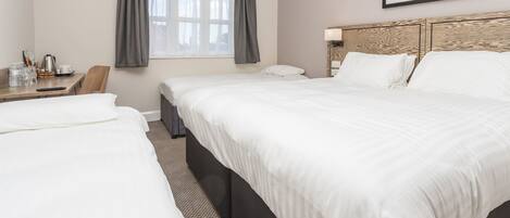A hotel room with two single beds, a wooden headboard, a desk with a chair, a window with curtains, and a framed picture on the wall.