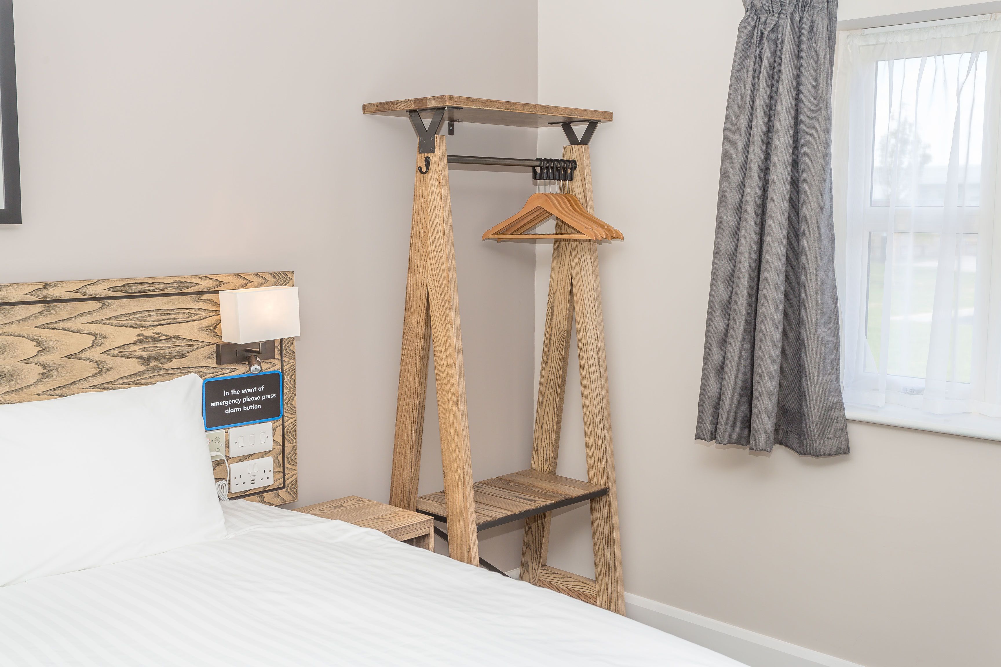 A bedroom with a bed, a wooden bedside table, a wall-mounted shelf with a hanging garment, and a window with curtains.