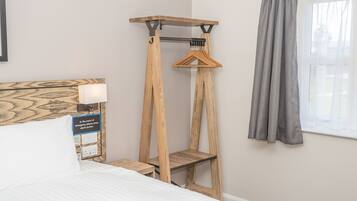 A bedroom with a bed, a wooden bedside table, a wall-mounted shelf with a hanging garment, and a window with curtains.