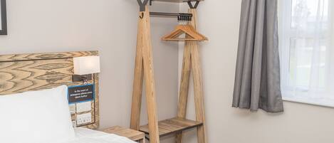 A bedroom with a bed, a wooden bedside table, a wall-mounted shelf with a hanging garment, and a window with curtains.