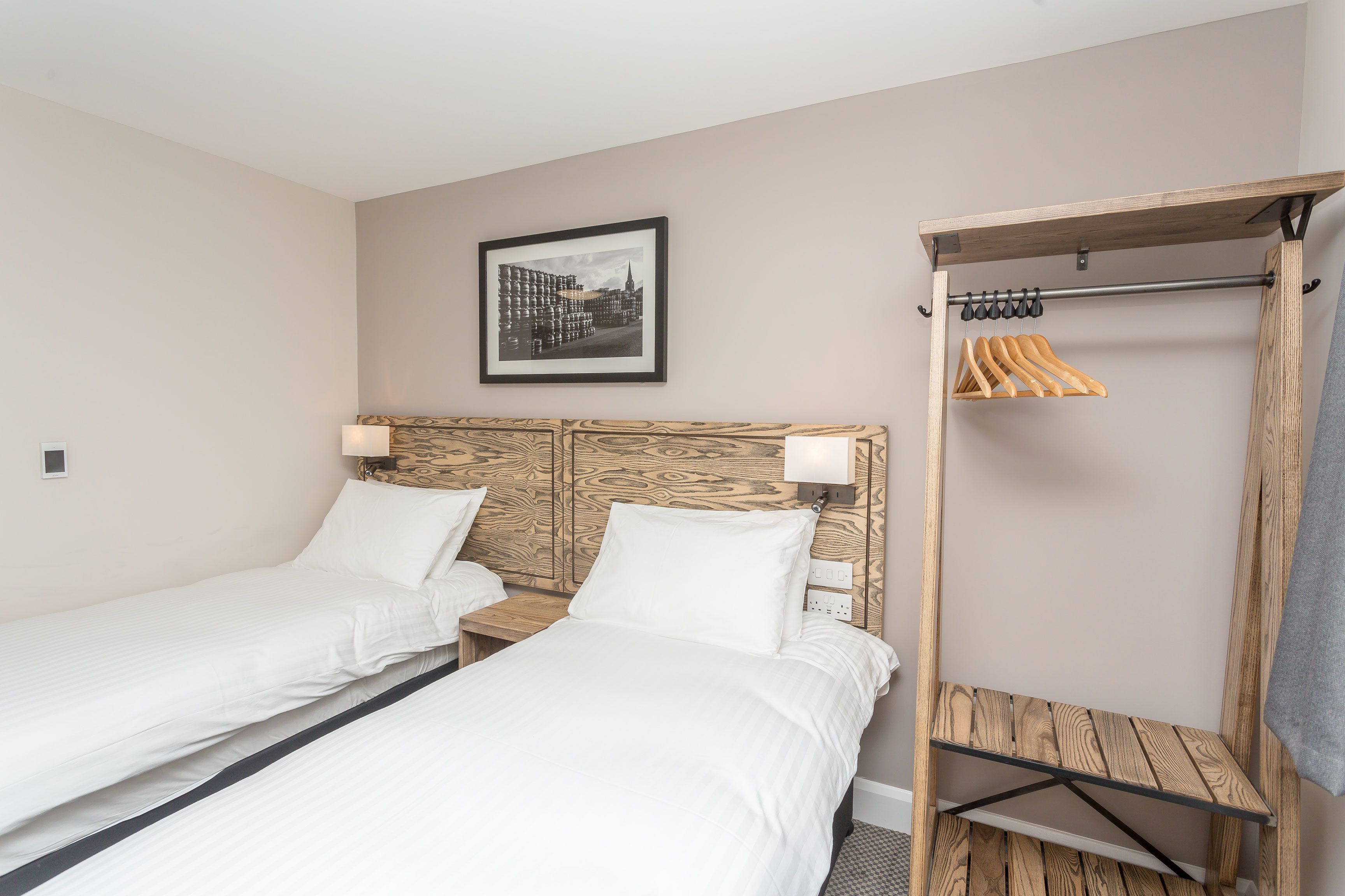 A hotel room with two single beds, a wooden headboard, a framed picture on the wall, and a clothing rack with hangers.