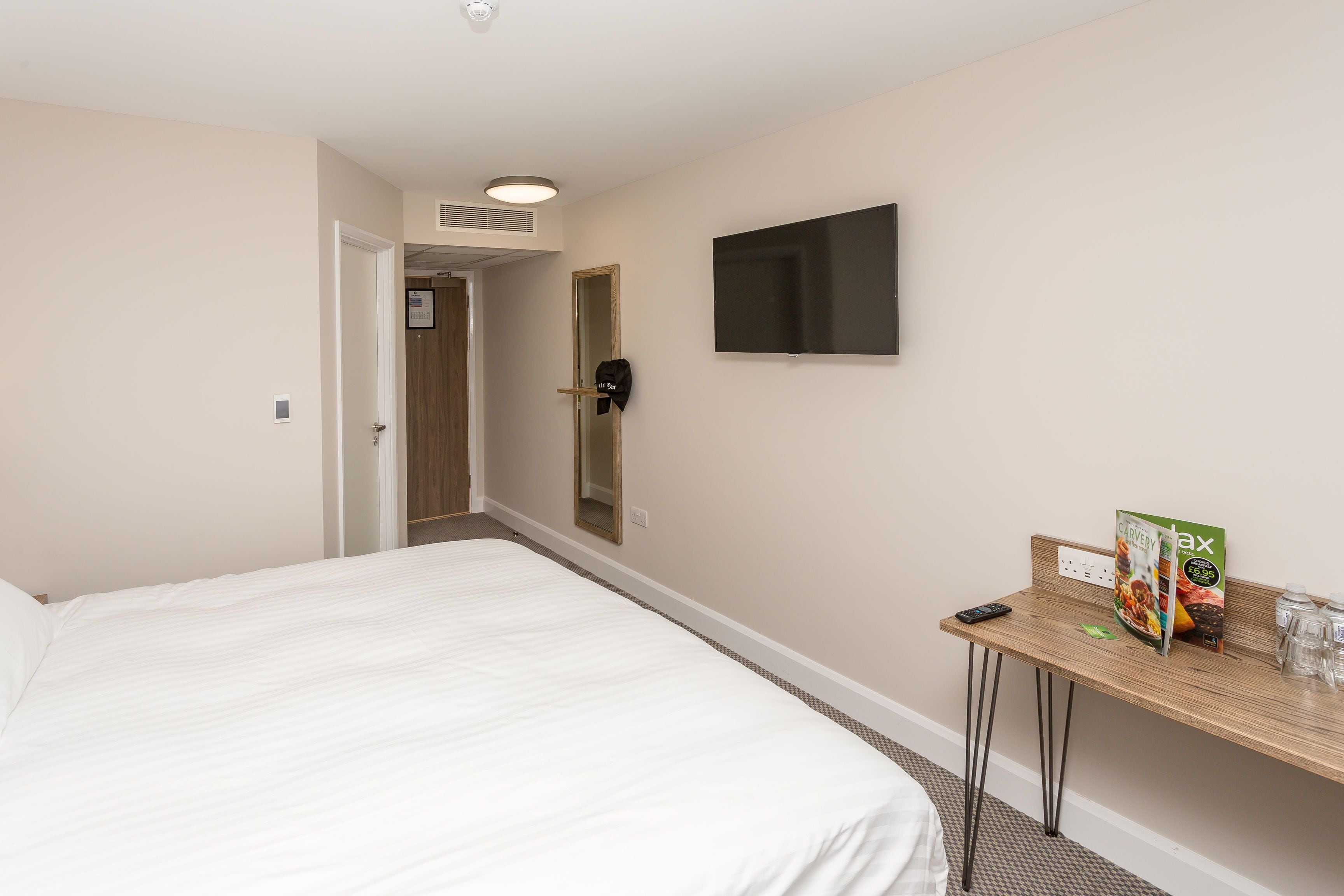 Una habitación de hotel con una cama, un televisor montado en la pared, una mesita con un control remoto, una revista y un vaso de agua.