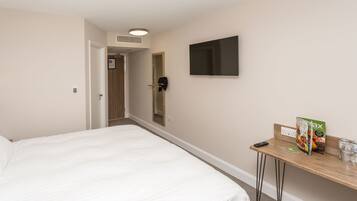 Una habitación de hotel con una cama, un televisor montado en la pared, una mesita con un control remoto, una revista y un vaso de agua.