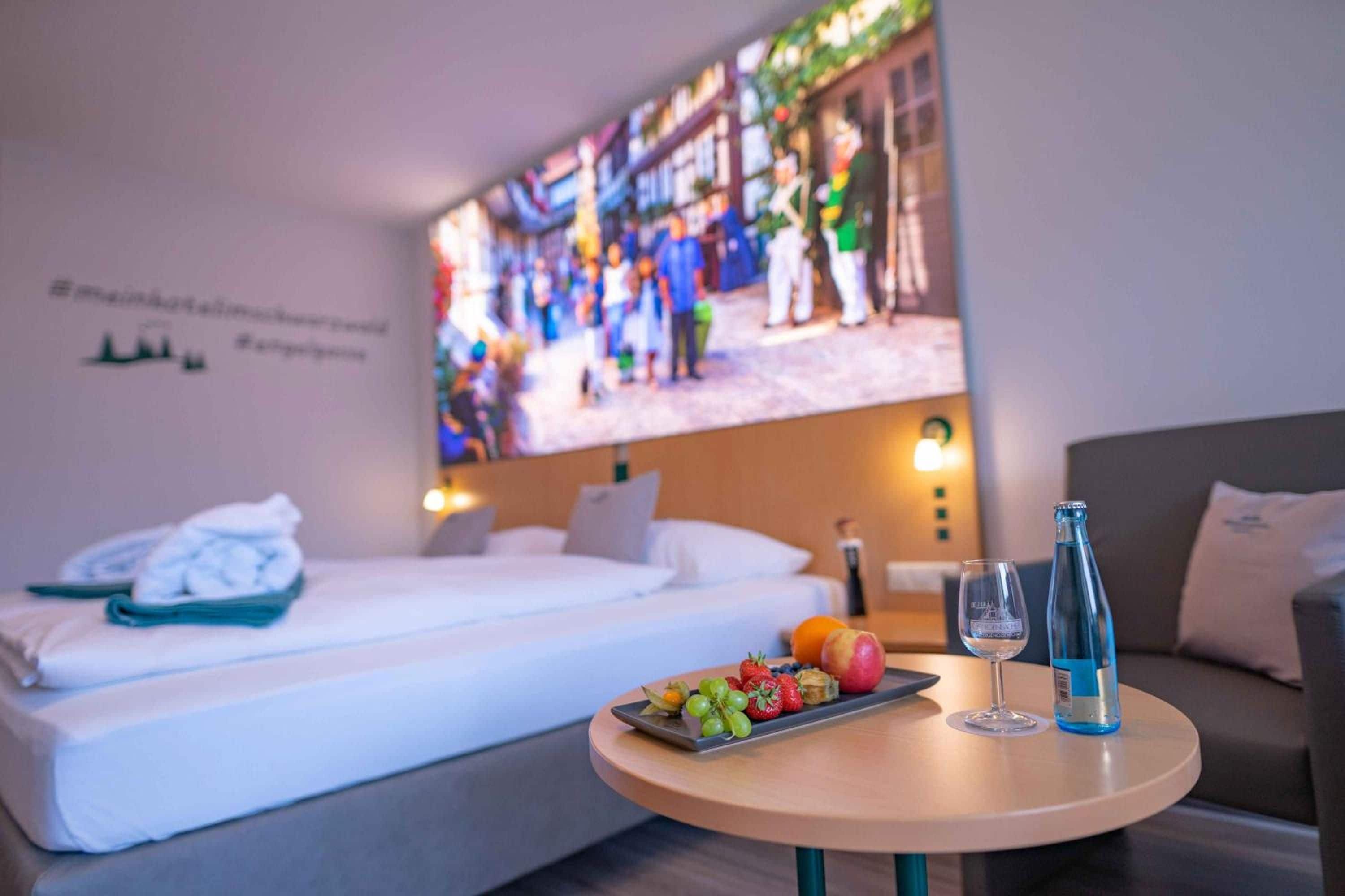 A hotel room with a bed, a small table with fruit and drinks, and a large screen displaying a busy street scene.
