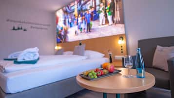 A hotel room with a bed, a small table with fruit and drinks, and a large screen displaying a busy street scene.