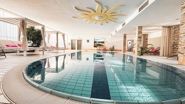 A large indoor swimming pool with a sun-shaped ceiling decoration, surrounded by lounge areas with cushioned chairs and a potted plant.