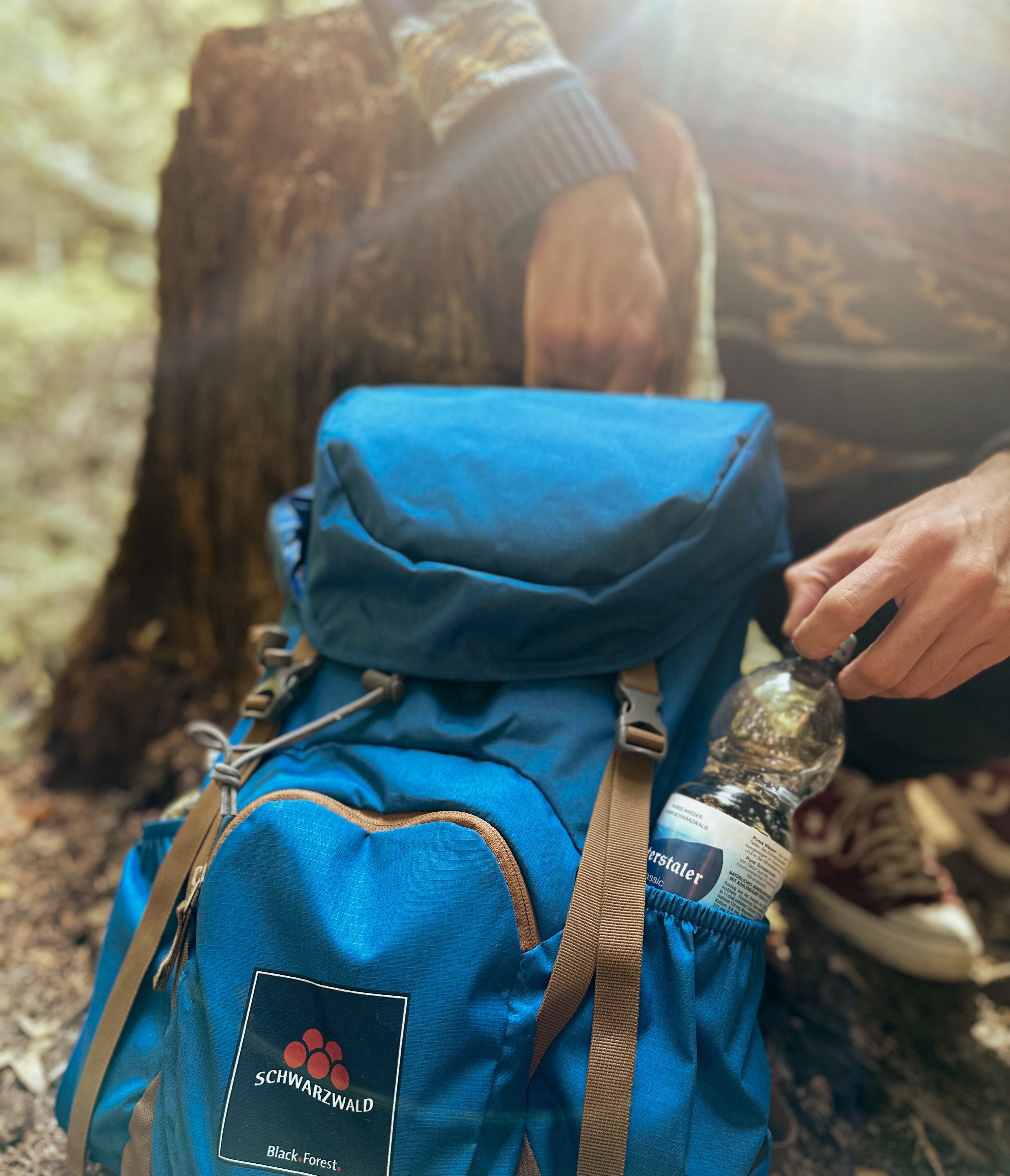 Persona con una mochila azul con la etiqueta 'SCHWARZWALD' y 'Selva Negra' sentada al aire libre, sosteniendo una botella.