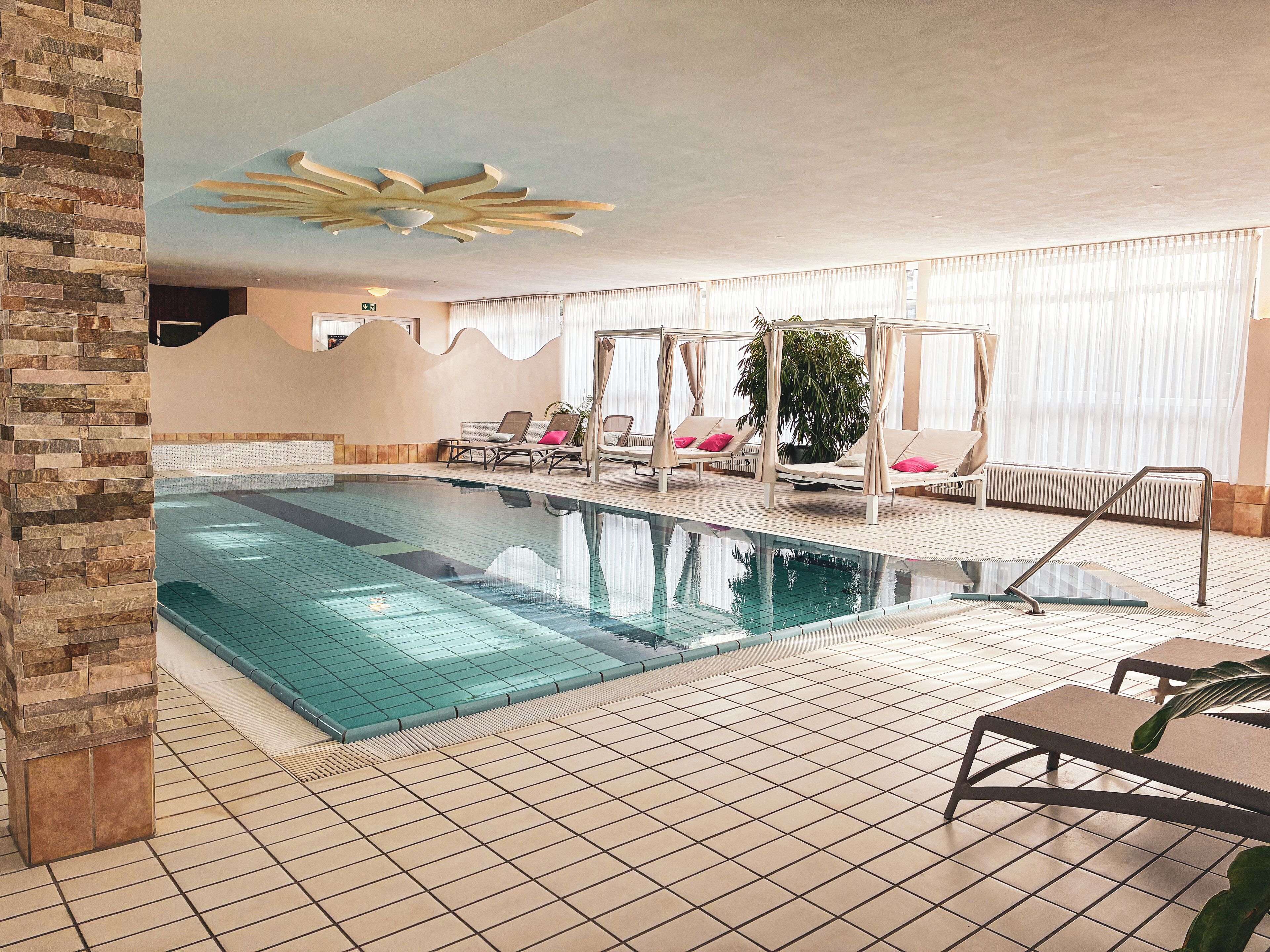An indoor swimming pool with tiled flooring, lounge chairs, and a ceiling with a sun-like design.