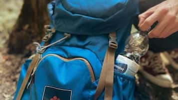 Uma pessoa com mochila azul, com logotipo e texto, sentada ao ar livre, com um toco de árvore ao fundo.