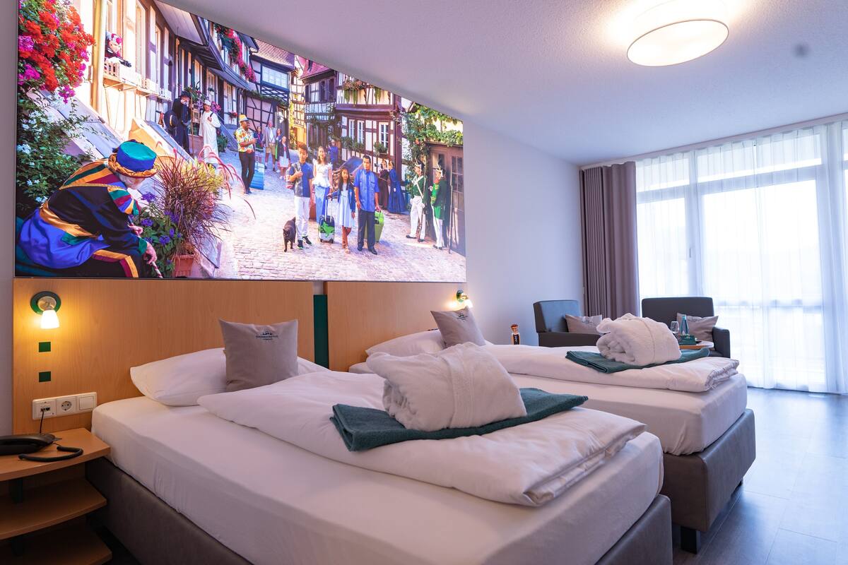 Habitación de hotel con dos camas, un televisor mostrando una escena callejera, un escritorio con un teléfono y una ventana con cortinas.