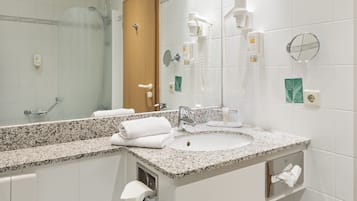 A hotel bathroom with a granite countertop sink, a mirror, a towel rack, a hand dryer, and a shower with a glass door.
