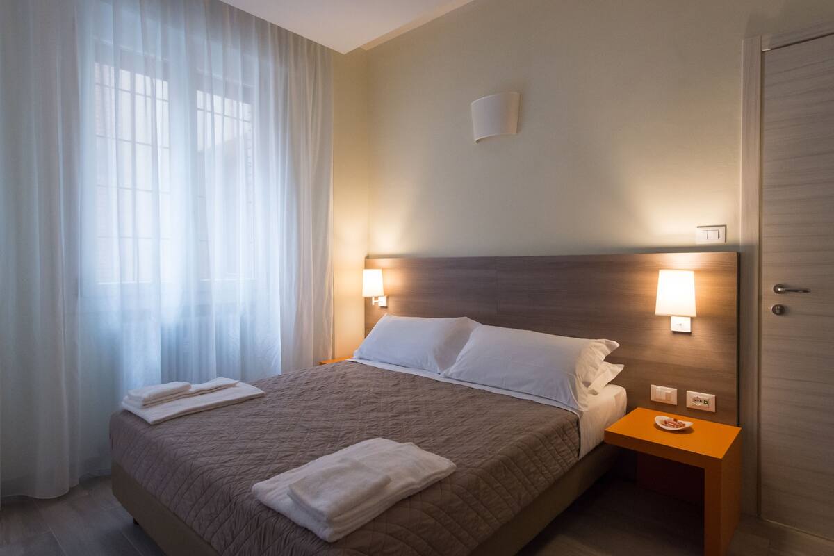 A neatly made bed with white linens, a bedside table with a plate, and a wall-mounted lamp.