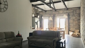 A living room with a sofa, a coffee table, and a dining area with chairs and a table. There is a stone wall and a clock on the wall.