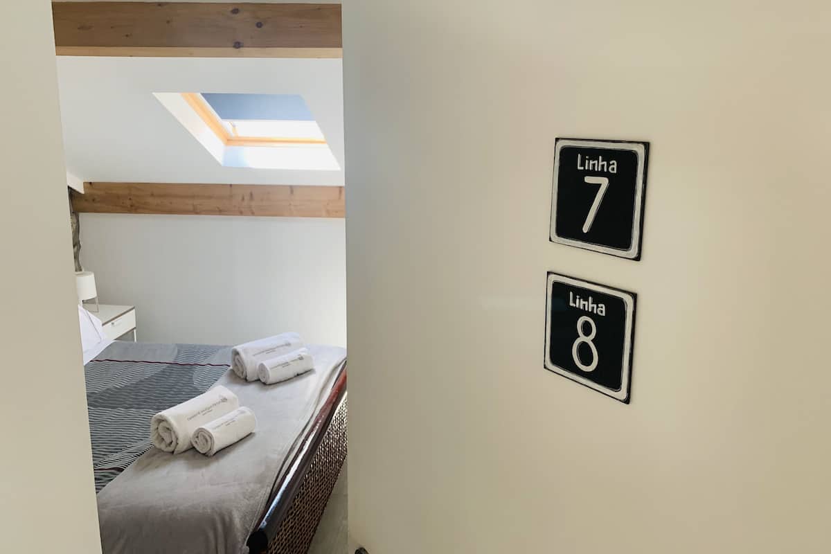 A hotel room with a bed, two wall-mounted signs reading "Linha 7" and "Linha 8", a skylight, and a small bedside table with a lamp.