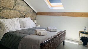 A bedroom with a stone wall, a sloped ceiling with a skylight, a bed with white linens, and a bedside table with a lamp and a bottle.
