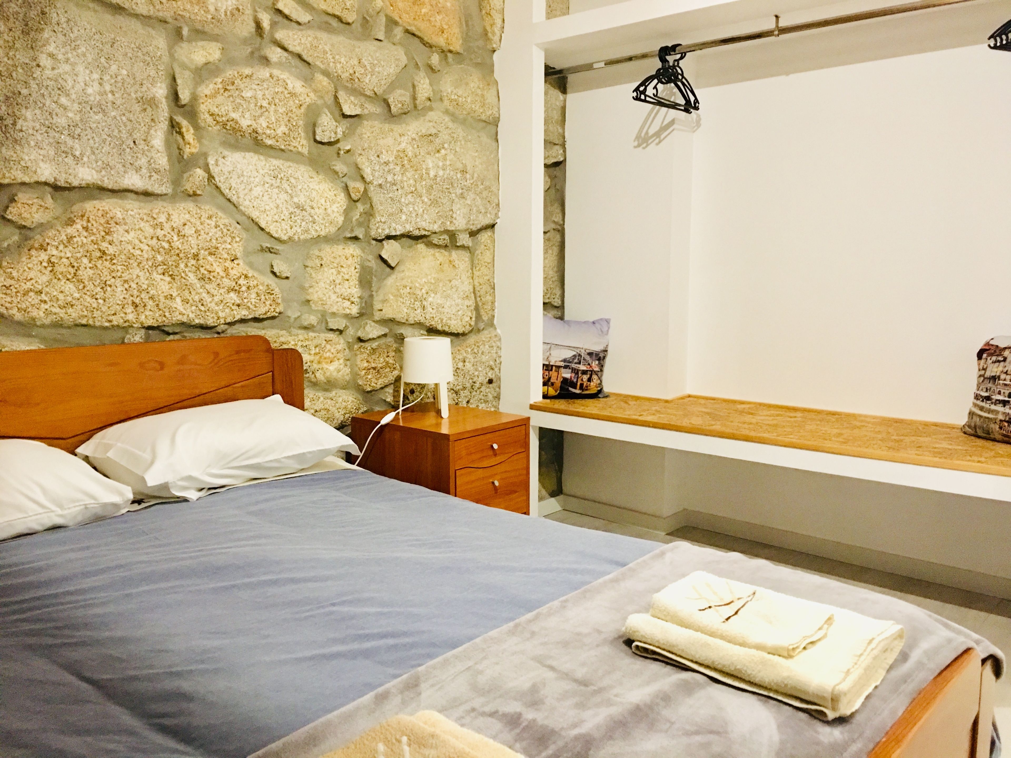 Un dormitorio con pared de piedra, cama de madera, mesita de noche y estante con una toalla y un bolso.