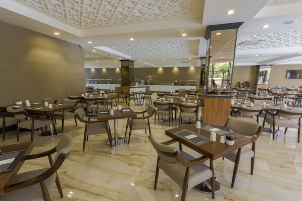 Un amplio comedor de hotel con varias mesas y sillas, un bar central y un diseño de techo moderno.