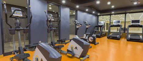 Un gimnasio con diversas máquinas de ejercicio, incluyendo cintas de correr y elípticas, dispuesto en un espacio amplio con suelo naranja y ventanas grandes.