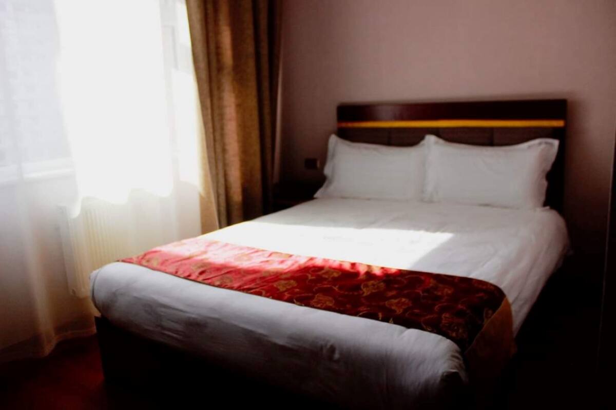 A hotel room with a neatly made bed, a window with curtains, and a radiator.