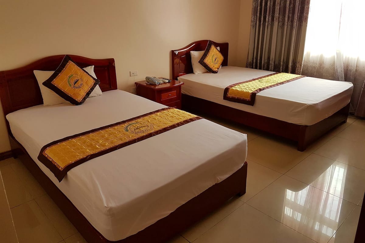 Two double beds with wooden headboards and matching decorative pillows, a bedside table with a phone, and a window with curtains.