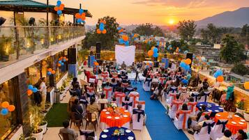 Un evento al aire libre con mesas y sillas dispuestas para los asistentes, un escenario para oradores y una vista panorámica al atardecer.