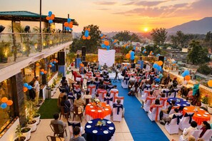 An outdoor event with tables and chairs arranged for guests, a stage for speakers, and a scenic sunset view. - Indira Nikunj Rubystone Exotic (Narendranagar)