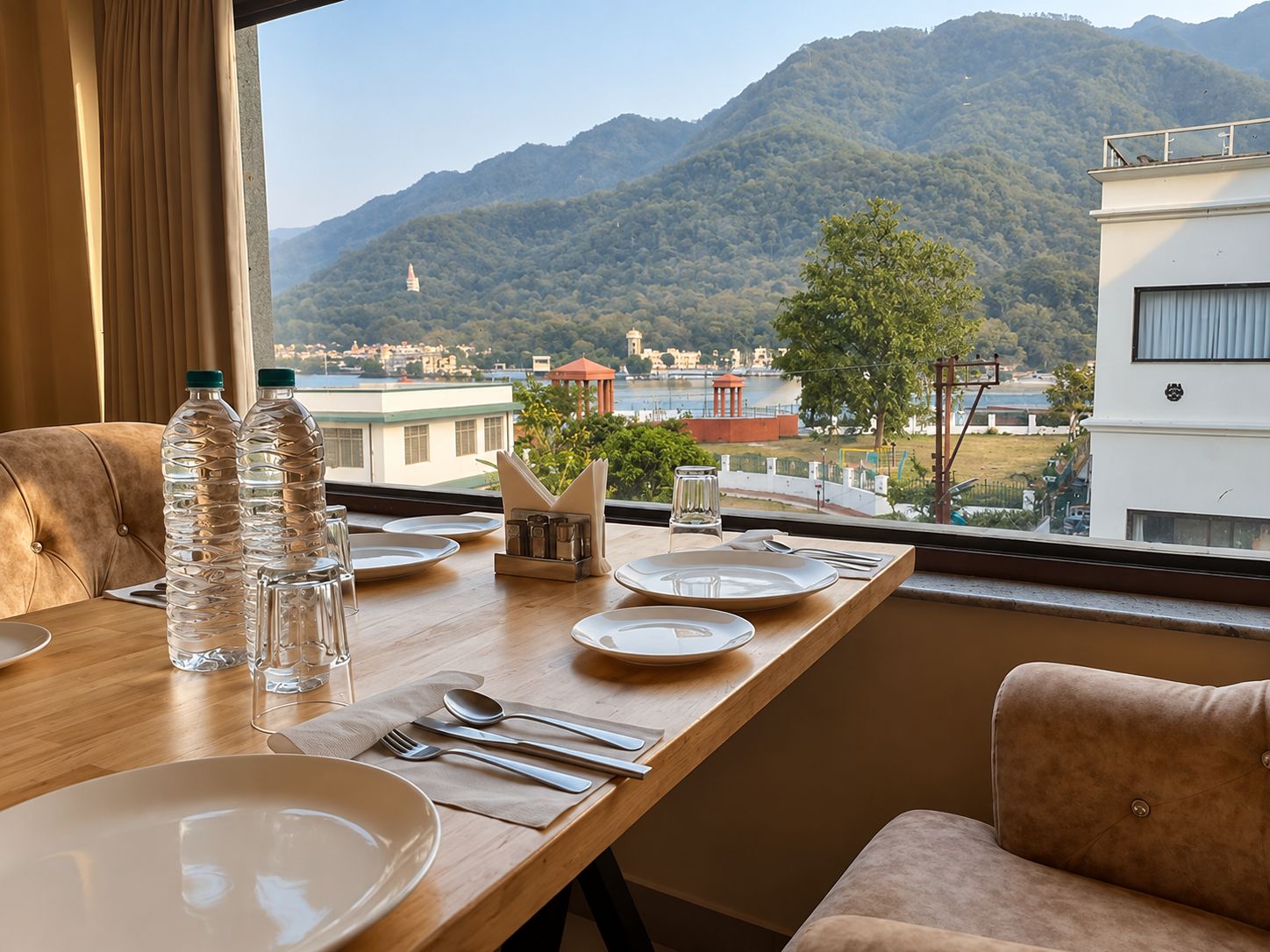 Mesa de comedor dispuesta con platos, cubiertos y botellas de agua, con vistas a un paisaje montañoso con edificios y un pabellón.