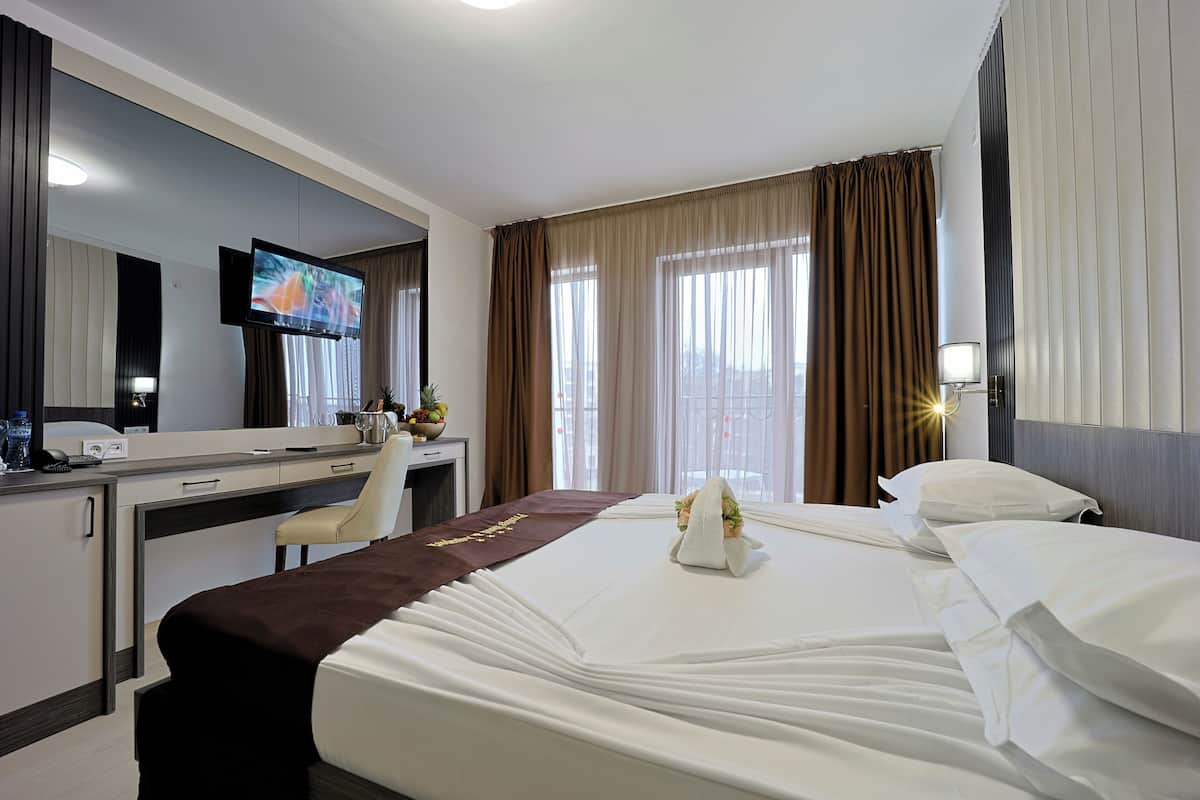 A hotel room with a bed, a desk, a television, a window with curtains, and a nightstand with a lamp.