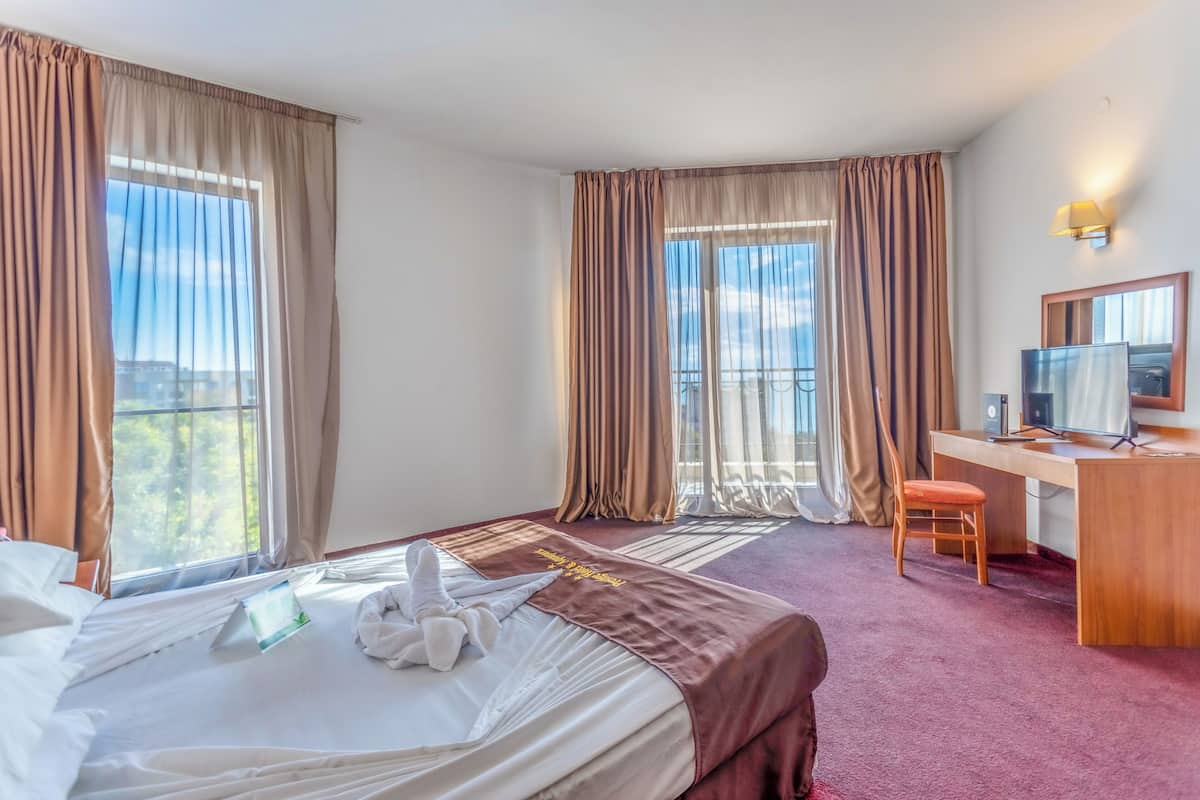 A hotel room with a bed, desk, chair, and large windows with curtains.