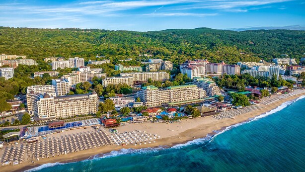 A coastal hotel with multiple buildings, a beach with sun loungers, and a view of the sea and hills. - Prestige Hotel and Aquapark (Golden Sands)