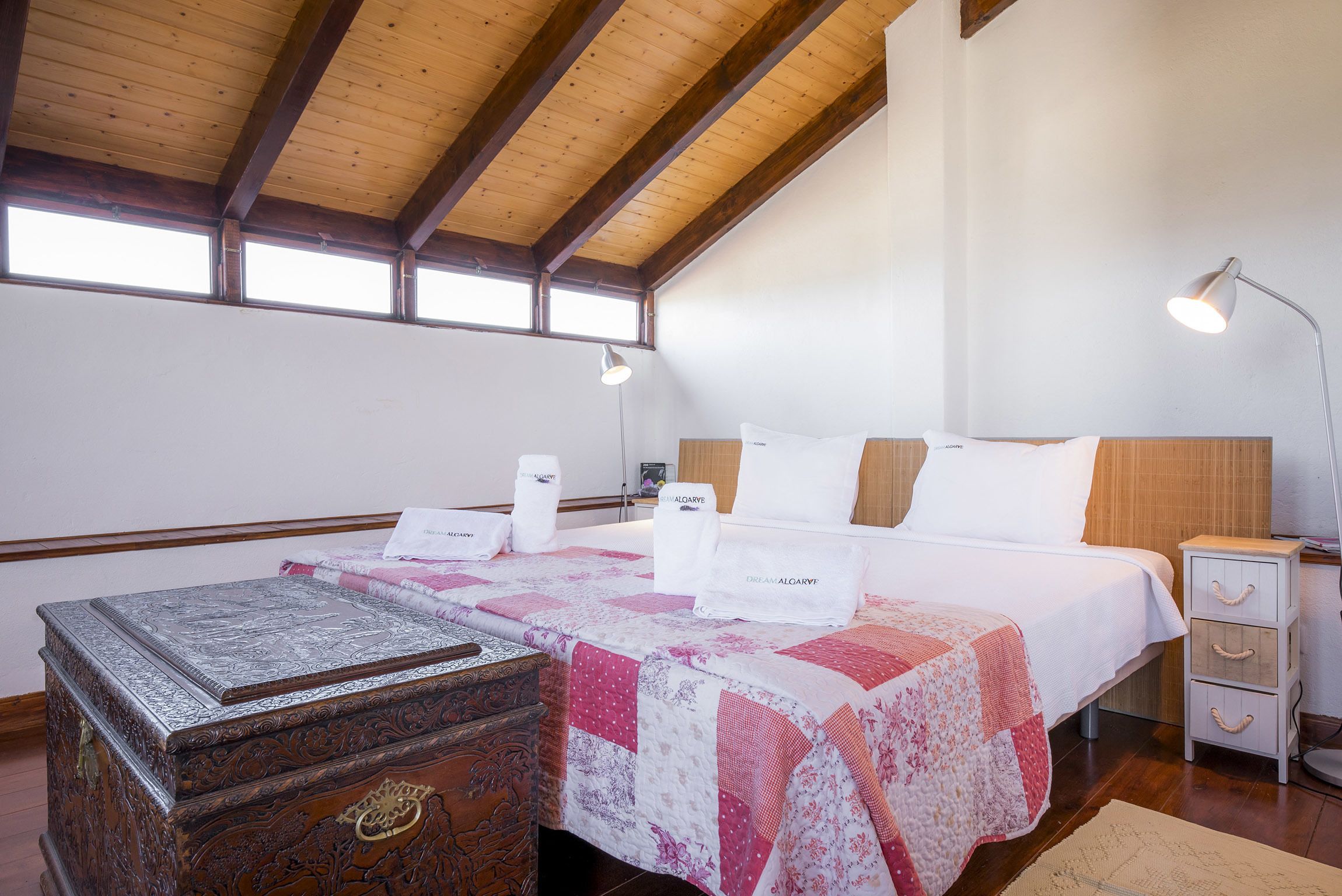 A bedroom with a wooden ceiling, a bed with a quilt, bedside lamps, a nightstand, and a decorative chest.