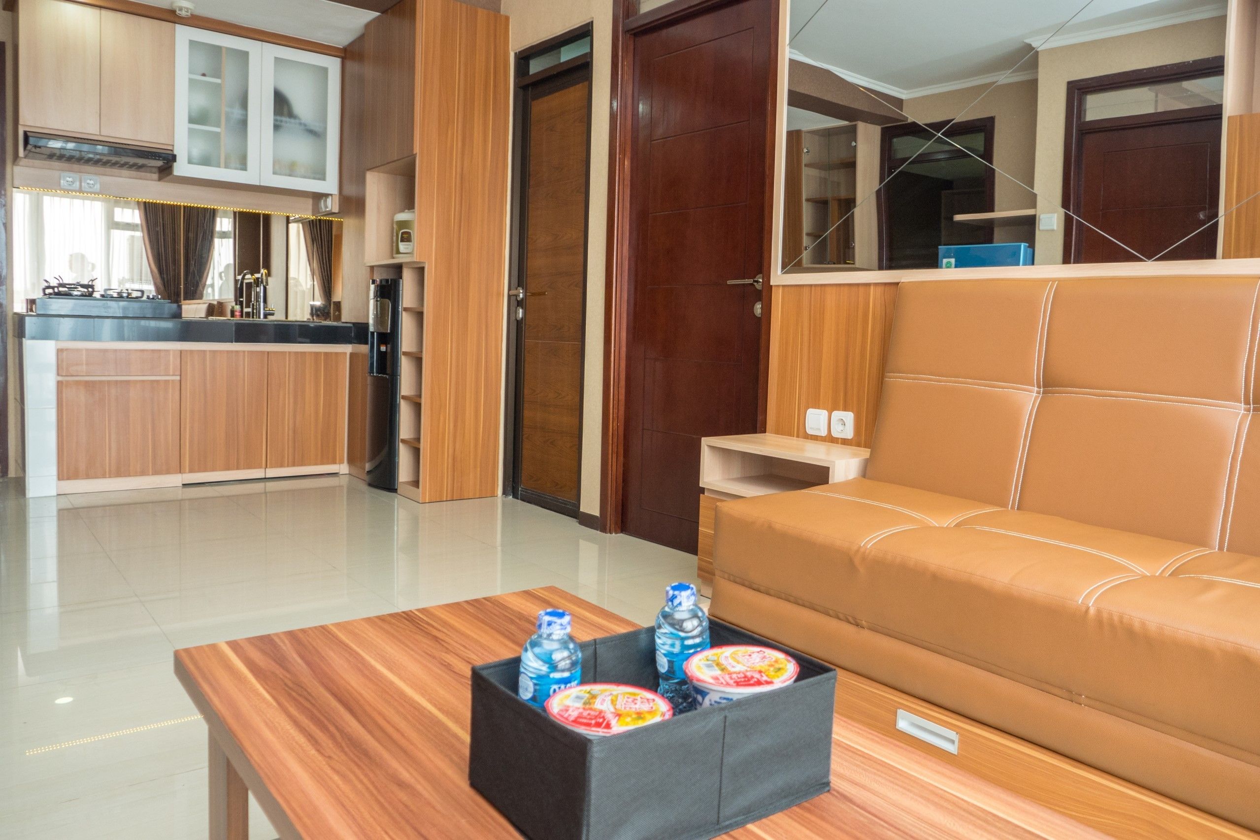 Una sala de estar moderna con un sofá, una mesa de centro con botellas de agua y una cocina con armarios de madera y un refrigerador.