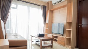 A modern living room with a sofa, a coffee table, a TV on a wooden stand, and a door to the right. - 3BR with Sofa bed at Gateway Pasteur Apartment (Cimahi)