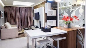 A modern living room with a white sofa, a dining table, and a TV. There are decorative shelves and a vase with flowers. - Furnished 1BR City View Casa Grande Apartment (Jakarta)