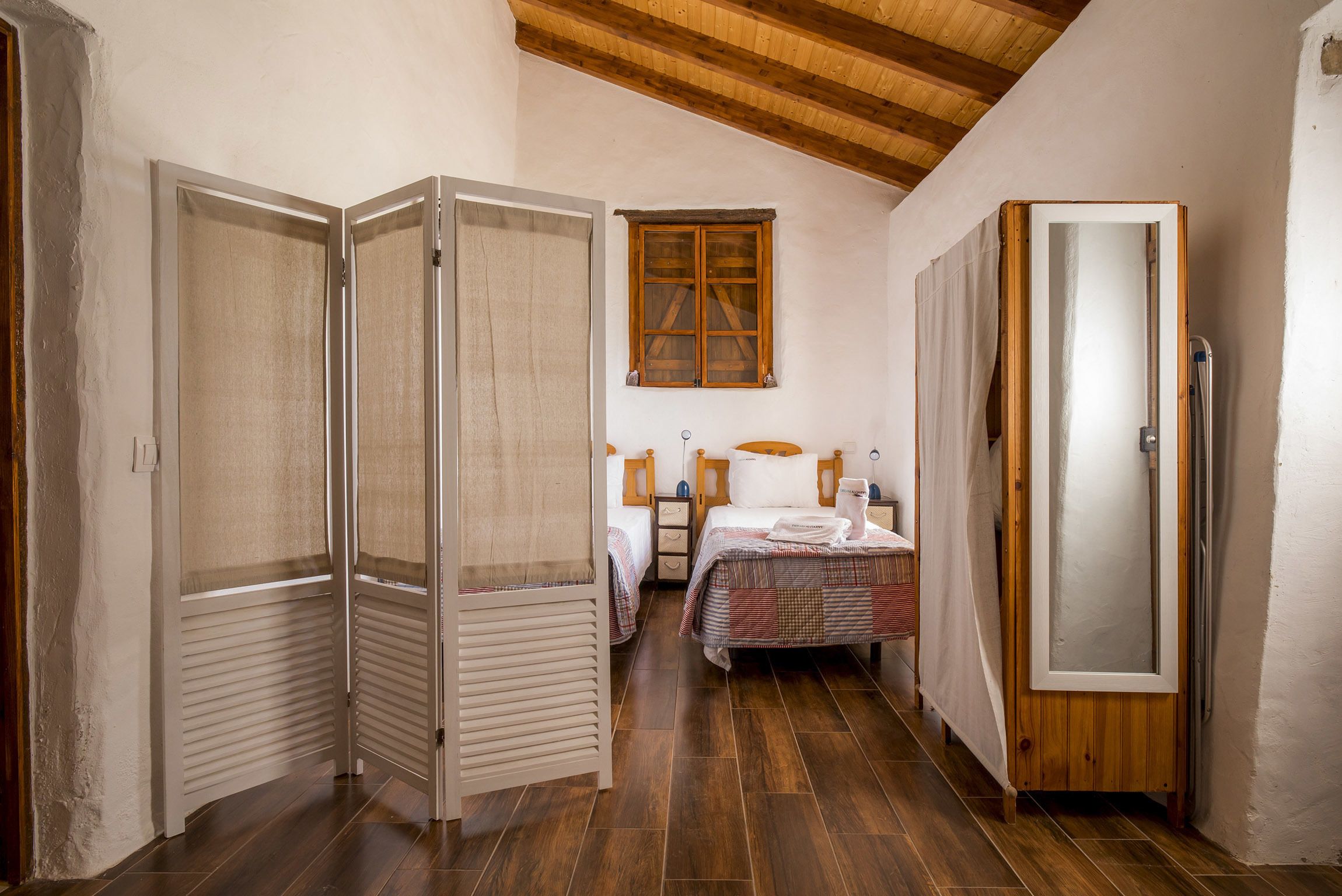A narrow room with a wooden ceiling, a bed with a checkered bedspread, a wooden wardrobe, and a window with wooden shutters.